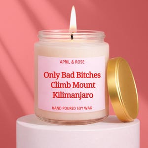 May include: A lit candle in a glass jar with a gold lid. The label reads "APRIL & ROSE" and "Only Bad Bitches Climb Mount Kilimanjaro. HAND POURED SOY WAX". The candle sits on a white pedestal against a pink background.