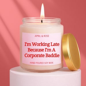 May include: A lit candle in a clear glass jar with a gold lid. The label reads "APRIL & ROSE" and "I'm Working Late Because I'm A Corporate Baddie. HAND POURED SOY WAX" on a pink background. The candle sits on a white pedestal.
