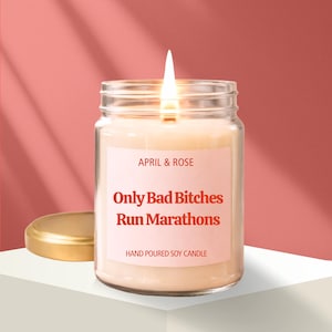 May include: A lit soy candle in a clear glass jar with a gold lid. The candle has a pink label with the text "APRIL & ROSE" and "Only Bad Bitches Run Marathons". The words "HAND POURED SOY CANDLE" are printed at the bottom of the label.