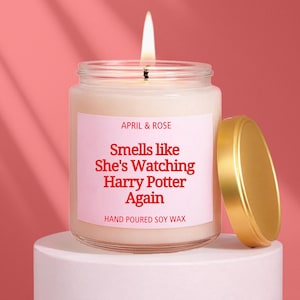 Puede incluir: Una vela encendida en un tarro de cristal con tapa dorada. La etiqueta dice "April & Rose" y "Smells like She's Watching Harry Potter Again". La vela es de cera de soja vertida a mano y está sobre un pedestal blanco.