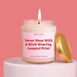May include: A clear glass jar candle with a lit wick and a gold lid. The label is light pink with red text that reads: APRIL & ROSE, Never Mess With A Bitch Wearing Leopard Print, HAND POURED SOY WAX. The candle contains off-white soy wax.
