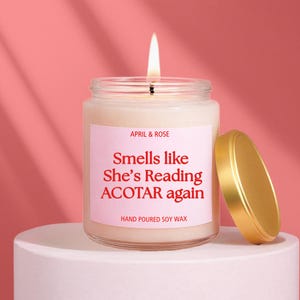 Könnte beinhalten: Eine brennende Kerze in einem durchsichtigen Glas mit einem goldenen Deckel. Auf dem Etikett steht "APRIL & ROSE" und "Smells like She's Reading ACOTAR again". Die Kerze steht auf einem weißen Podest vor einem rosa Hintergrund. Der Text "HAND POURED SOY WAX" steht ebenfalls auf dem Etikett.