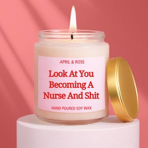 Könnte beinhalten: Eine brennende Kerze in einem durchsichtigen Glas mit goldenem Deckel. Das Etikett lautet "APRIL & ROSE Look At You Becoming A Nurse And Shit HAND POURED SOY WAX" auf rosa Hintergrund. Die Kerze steht auf einem weißen Sockel.