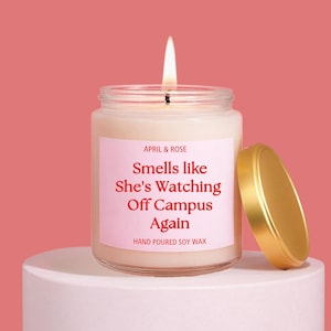 Puede incluir: Una vela encendida en un tarro de cristal transparente con tapa dorada. La etiqueta dice "APRIL & ROSE" y "Smells like She's Watching Off Campus Again". La vela está sobre un pedestal blanco sobre un fondo rosa. Cera de soja vertida a mano.
