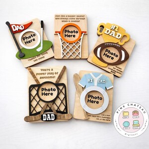 May include: A collection of wooden photo frames with sports themes, including golf, basketball, football, hockey, and baseball. Each frame has a cutout for a photo and the word "DAD" with various phrases. The frames are light brown with colourful accents.