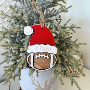 May include: A wooden Christmas bauble shaped like a football with a Santa hat on top. The bauble is decorated with a brown and white football design and the name "KHALED" is written on the hat. The year "2024" is written on a small white circle attached to the hat.
