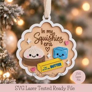 May include: A scalloped wooden ornament featuring the text In my Squishies era, a dumpling in a steamer, a blue cube, and a yellow stick of butter labeled 1 oz NET WT. 28g. The design includes small pink hearts and white four-pointed stars.