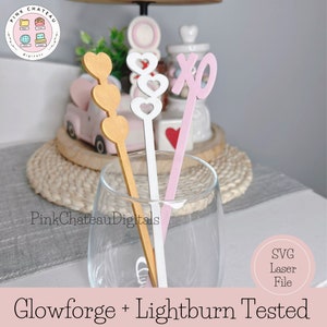Puede incluir: Tres palitos de madera para el Día de San Valentín con diferentes diseños. Un palito tiene tres corazones dorados, otro tiene tres corazones blancos y el último tiene un XO rosa. Los palitos están en un vaso transparente. Probado Glowforge + Lightburn. Archivo SVG Laser.