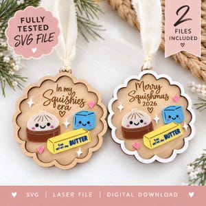 Pode incluir: Dois ornamentos redondos de madeira com bordas recortadas e fitas brancas. Cada um apresenta um bolinho, um cubo azul e um tablete de manteiga amarelo com o texto 1 oz NET WT. 28g BUTTER. Um diz In my Squishies era, o outro Merry Squishmas 2026.