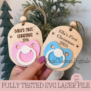Pode incluir: Enfeites de Natal de madeira em forma de chupeta, um rosa e outro azul, com os textos "Baby's First Christmas 2026" e "Ella's First Christmas 2025". Cada enfeite tem um cordão de juta. Corte a laser.
