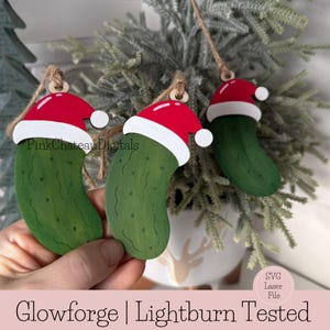 May include: Three wooden Christmas ornaments shaped like pickles, each wearing a red Santa hat with a white trim and a white pom-pom. The ornaments are suspended by twine and are set against a backdrop of evergreen branches.