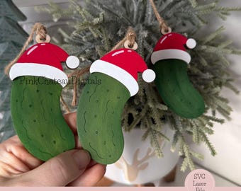 May include: Three wooden Christmas ornaments shaped like pickles, each wearing a red Santa hat with a white trim and a white pom-pom. The ornaments are suspended by twine and are set against a backdrop of evergreen branches.