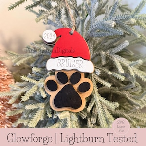 May include: A wooden Christmas bauble featuring a red Santa hat, a white paw print, and the name "BRUISER" inscribed on it. The bauble also has a small circle with the year "2024" written on it.