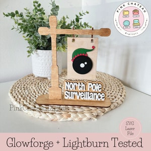 以下が含まれることがあります： 「北極監視」という文字と、黒い目をした漫画の妖精が描かれた木製の看板。看板は木製の街灯の柱に取り付けられています。看板はレーザーカット用に設計されています。