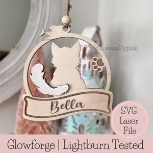 Pode incluir: Um enfeite de Natal de madeira com a silhueta de um gato com uma cauda branca, uma pegada e uma faixa com o nome "Bella". O enfeite é projetado para corte a laser e é testado para uso com Glowforge e Lightburn.