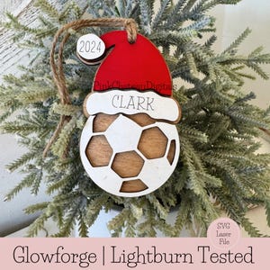 May include: A wooden Christmas bauble shaped like a football with a Santa hat on top. The bauble is white with brown details and the name "CLARK" is written on the hat. The year "2024" is written on a small tag attached to the bauble.