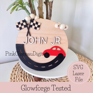 May include: A wooden, circular sign with the name "JOHN JR" in white letters. A red car is on a black road, and two checkered flags are at the top. The sign has a light wood finish and is decorated with wooden beads.