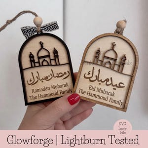 Op de afbeelding: Twee houten Ramadan- en Eid Mubarak-ornamenten. Elk ornament heeft een moskeesilhouet en Arabische kalligrafie. Het Ramadan-ornament heeft een zwarte rand en een zwart-wit lint. Beide zeggen "The Hammoud Family".