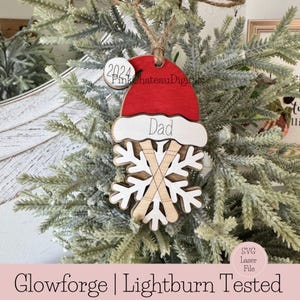 May include: A wooden Christmas bauble featuring a red Santa hat, a snowflake design, and the word "Dad" inscribed on it. The bauble is hanging on a Christmas tree.