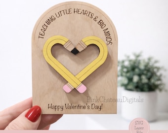 May include: A wooden Valentine's Day card with a heart-shaped pencil design. The pencils are yellow with pink erasers and a black tip. The card reads "Teaching Little Hearts & Big Minds" and "Happy Valentine's Day!"