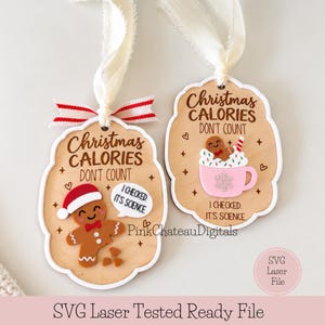 May include: Two wooden Christmas ornaments with white ribbon hangers. One features a gingerbread person in a Santa hat with the text Christmas Calories Don't Count I Checked It's Science. The other shows a gingerbread person in a pink mug.