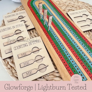Puede incluir: Un tablero de juego de madera con una pista para jugar un juego de conteo. El tablero está hecho de madera clara y tiene rayas rojas, verdes y azules. Hay cuatro piezas de madera con las palabras "Counting in Style!" e iniciales en ellas. Las iniciales son GR, JG y MK. El tablero de juego está etiquetado como "SVG Laser File".