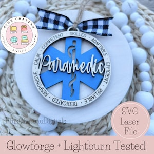 Może przedstawiać: Okrągła, biała drewniana ozdoba z niebieskim symbolem ratownika medycznego i napisem "Paramedic" w białym skrypcie. Ozdoba ma czarno-białą kratowaną wstążkę i słowa "Kompetentny, Wykwalifikowany, Elastyczny, Wydajny, Niezawodny, Oddany, Reagujący, Spokojny" na krawędzi.