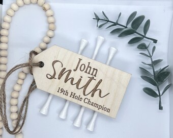 May include: A wooden tag with the text "John Smith 19th Hole Champion" engraved on it. Four white golf tees are attached to the tag with twine.