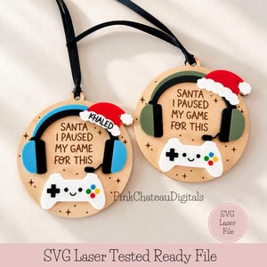 Op de afbeelding: Twee ronde houten ornamenten met zwarte linten. Elk item bevat een witte gamecontroller, een kerstmuts en een koptelefoon. De tekst luidt: SANTA I PAUSED MY GAME FOR THIS. Eén muts is gepersonaliseerd met de naam KHALED. Kleine sterren zijn gegraveerd.