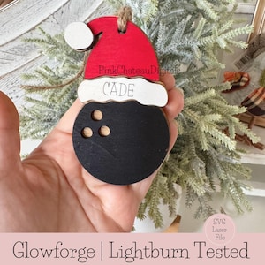May include: A wooden Christmas bauble shaped like a bowling ball with a Santa hat on top. The hat is red with white trim and the name "CADE" is written on the white trim. The bowling ball is black with white pin holes. The ornament is designed for use with a Glowforge or Lightburn.