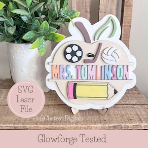 May include: A wooden apple-shaped teacher appreciation sign with the text "MRS. TOMLINSON" in rainbow colors. The apple has a green leaf, a pink and yellow pencil, a soccer ball, a volleyball, and a hockey stick. The sign is on a wooden surface.