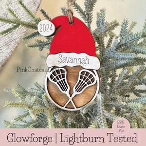 May include: A wooden Christmas bauble with a red Santa hat, the name "Savannah" and the year "2024" on it. The bauble features two lacrosse sticks crossed in the centre.