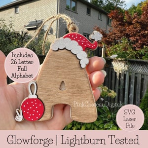 May include: Wooden Christmas ornament shaped like the letter A, decorated with a red Santa hat and a red bauble with white polka dots. The ornament is held in a hand. The text "Includes 26 Letter Full Alphabet" is visible in the image. The text "Pink Chateau Digitals" is visible in the image. The text "SVG Laser File" is visible in the image. The text "Glowforge | Lightburn Tested" is visible in the image.