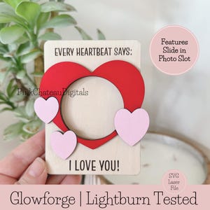 May include: A wooden heart-shaped photo frame with a red heart cutout and three pink hearts. The text "EVERY HEARTBEAT SAYS: I LOVE YOU!" is displayed. The frame has a slot for a photo. The words "Glowforge | Lightburn Tested" are also visible.