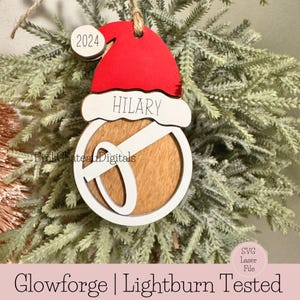 May include: A wooden Christmas bauble with a red Santa hat, a white name tag that says "HILARY", and a white letter "O" on a brown background. The ornament also has a small white circle with the year "2024" on it.