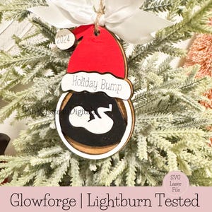 May include: A wooden Christmas bauble featuring a red Santa hat and the text "Holiday Bump". The bauble is decorated with a white silhouette of a baby inside a black circle. The bauble is hanging on a green Christmas tree.