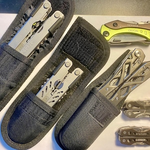 May include: Five multi-tool pocket knives in various colors and styles, including a black and silver Gerber multi-tool, a green and silver Gerber Crucial multi-tool, and a black and silver Leatherman multi-tool. The multi-tools are displayed in black nylon pouches.