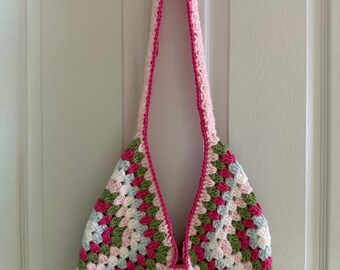 Puede incluir: Bolso tote de crochet hecho a mano con un patrón de cuadrados de la abuela. El bolso presenta una mezcla de cuadrados rosas, verdes, blancos y celeste. La correa y el ribete son de color rosa liso. El bolso está colgado de un gancho negro.