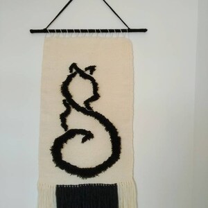 Puede incluir: Un tapiz de pared tejido blanco y negro con una silueta de gato negro. El tapiz tiene un borde de flecos.