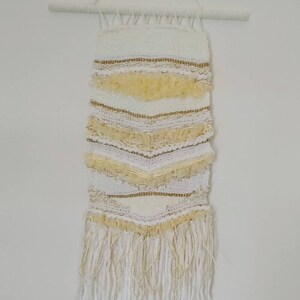 Puede incluir: Un tapiz de pared tejido blanco y dorado con un borde de flecos. El tapiz está hecho con diferentes texturas y patrones de hilo.
