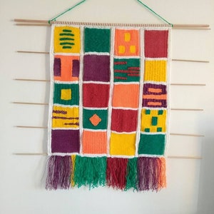 Puede incluir: Un tapiz de pared de punto de colores con un patrón geométrico. El tapiz está formado por cuadrados en tonos de rojo, naranja, amarillo, verde y morado. Los cuadrados están dispuestos en una cuadrícula y tienen diferentes formas geométricas en su interior. El tapiz tiene un borde de flecos.