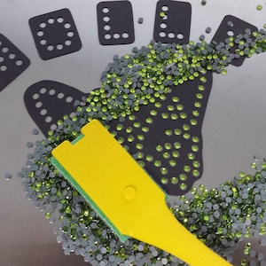 May include: A yellow and green tool for picking up small rhinestones, scattered on a silver surface, with black stencils in the background.