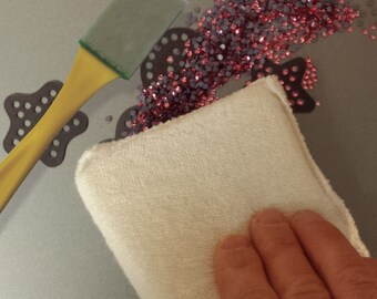 May include: A close-up of a white microfiber cloth being used to clean up pink and silver rhinestones scattered on a gray surface. A yellow tool with a green handle is also visible in the image.