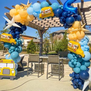 May include: A balloon arch decorated with yellow school bus balloons, blue and white star balloons, and blue and yellow balloons. The arch is set up outdoors against a backdrop of a wooden pergola and a blue pool.