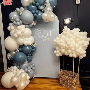 May include: A balloon arch in shades of white, gray, and blue, adorned with small white flowers, frames a light blue backdrop with the text "On Cloud Nine." A cloud-shaped balloon arrangement sits atop a woven basket, resembling a hot air balloon.