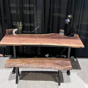 Puede incluir: Un escritorio de nogal de borde vivo con un marco de metal negro y un banco de nogal de borde vivo a juego. El escritorio tiene una regleta de enchufes integrada y una lámpara negra en el lado derecho.