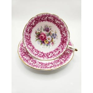 Puede incluir: Un juego de taza y plato de porcelana con un vibrante patrón floral de color magenta en el borde y un ramo central de flores coloridas que incluye rosas y margaritas. Los bordes de la taza y el plato están rematados con una fina línea dorada.