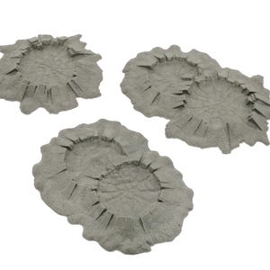 May include: Three gray, crater-shaped terrain pieces. Each piece has a circular depression in the center with jagged edges and radial lines. The pieces are made of a textured material and are designed for tabletop gaming.