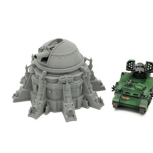 Op de afbeelding: Grijs, gedetailleerd model van een koepelstructuur met een groot kanon en een groen militair voertuig met raketwerpers. Modellen voor tabletop games of modelbouw.