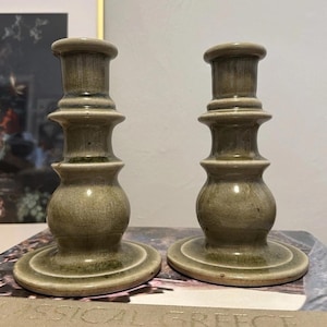 May include: Two olive green ceramic candlesticks with a bulbous body and tiered design. The candlesticks have a round base and a top designed to hold a candle. The candlesticks are displayed on a book.